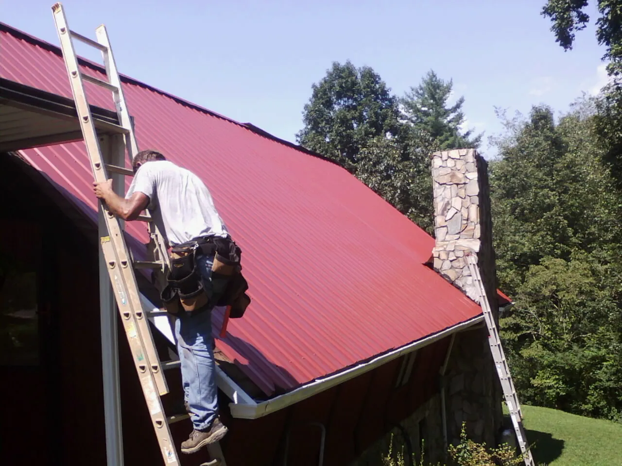 Roofer on a ladder beside a red metal roof and stone chimney — Soffit & Fascia Repair by Roofing Brothers in Moclips